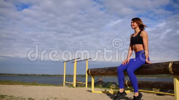 运动型深色头发的女孩坐在河边的一根木头上在力量训练前进行热身视频的预览图