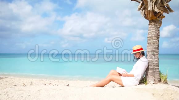 年轻女子坐在棕榈树下的白色海滩上看书视频的预览图