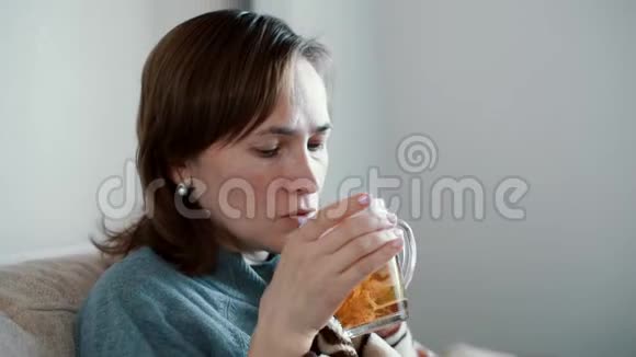 一个女人坐在家里的沙发上温暖起来裹着格子喝着热茶视频的预览图