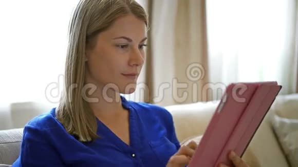 坐在沙发上的女人拿着她的平板电脑浏览互联网微笑与她的朋友交流视频的预览图
