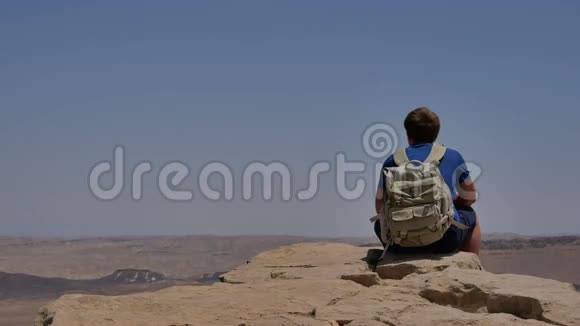 年轻男子背着背包坐在悬崖边享受沙漠景色视频的预览图