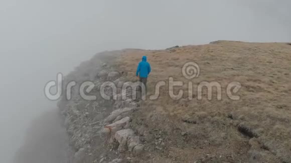 徒步旅行者穿着一件带背包的蓝色夹克站在云层上方陡峭的高原边缘视频的预览图