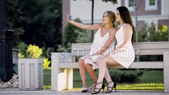 穿着夏季服装的可爱女孩和成年女孩在公园里自拍女士们在温暖的一天坐在长凳上视频的预览图