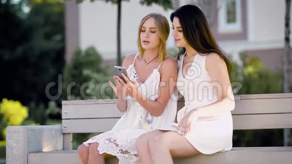 可爱的女孩给她的女朋友看了一张荧光照片她的手机上放着一个假期她们坐在公园的长凳上视频的预览图
