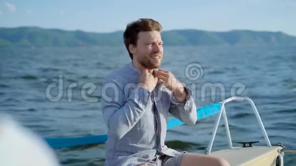 一个坐游艇的成年男子调整了他的衬衫那人从阳光下眯起眼睛阳光照耀着他视频的预览图