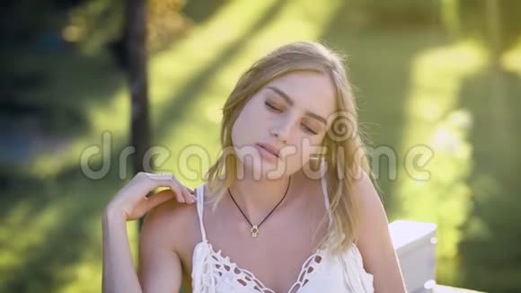 一位年轻可爱的女孩在夏天坐在外面的公园里的肖像这位女士嬉戏地抚摸着她的头发视频的预览图