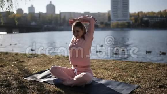 在城市河岸上穿着运动服的女孩在一个特殊的垫子上做瑜伽听音乐视频的预览图