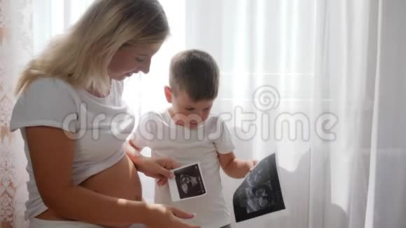 年轻的怀孕妈妈带着甜美的孩子在明亮的自然光线下在家里看摄影声像视频的预览图