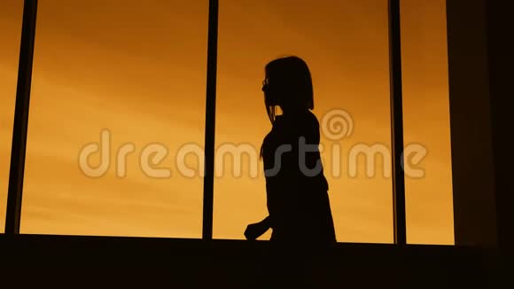 一位身材苗条的长发女子在暮色中沿着全景式的落地窗走着她的轮廓是黑暗的视频的预览图
