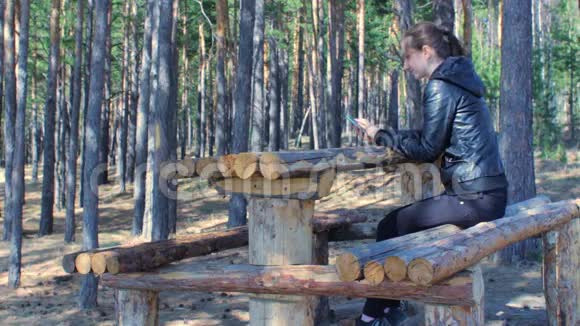 年轻女子坐在公园森林里的木凳上用电话秋天视频的预览图