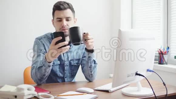 年轻的上班族在办公室里用手机坐在桌子旁拿着电脑电话喝着咖啡或茶视频的预览图