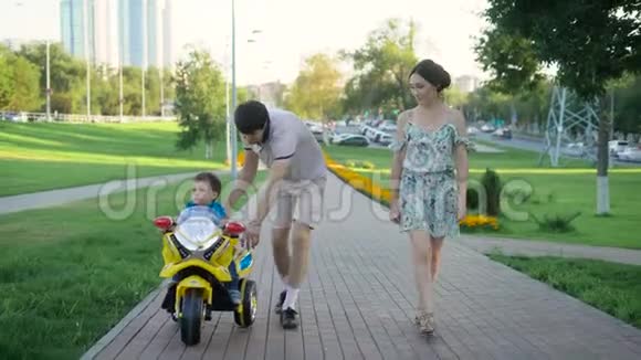夏天阳光明媚一家人在现代城市的小巷里散步父亲正在帮他的小儿子骑摩托车视频的预览图