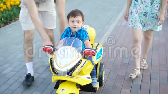 夏天小男孩在一条城市小巷里骑着一辆明亮的玩具摩托车父母陪着他视频的预览图
