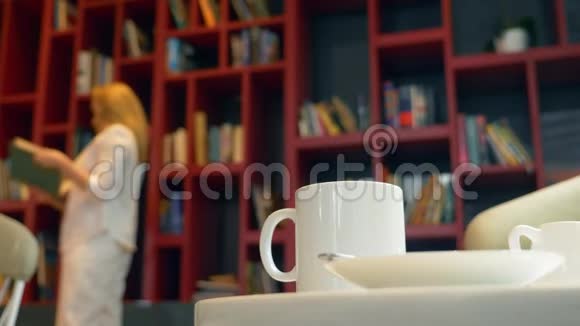 专注于一个杯子和一杯热饮料在图书馆的桌子上布鲁尔年轻的女人选择一本书来读站在里面视频的预览图