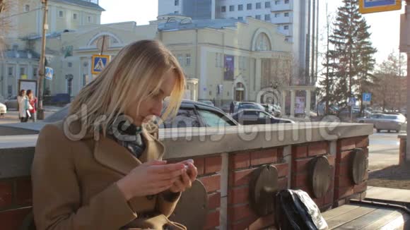 在阳光明媚的城市街道上坐在长凳上用智能手机看书的女人视频的预览图