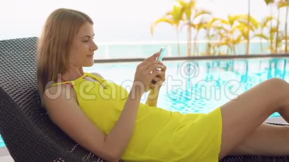 穿着黄色衣服的女人坐在靠近游泳池的日光浴床上用智能手机和朋友聊天视频的预览图
