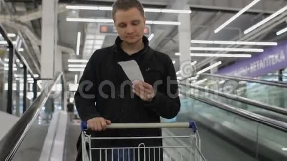 一位年轻的帅哥带着支票和手推车在购物中心的自动扶梯上旅行视频的预览图