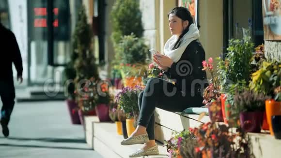 美丽的女人坐在城市街道的长凳上用她的智能手机视频的预览图