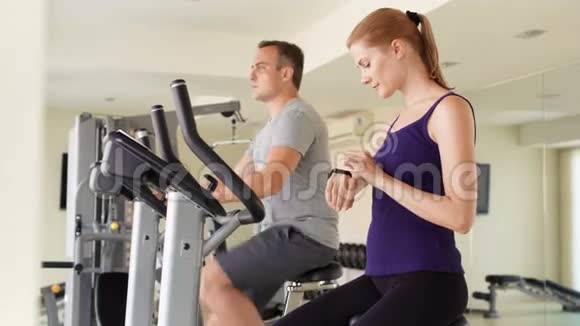 适合积极的夫妇在健身房锻炼自行车女人用智能手表检查生命体征视频的预览图