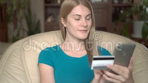 聪明漂亮的年轻女人坐在家里的椅子上用信用卡在线购买智能手机视频的预览图