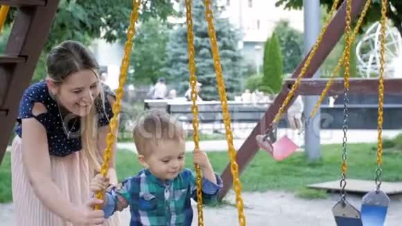 快乐的年轻母亲在公园里摇摆着蹒跚学步的儿子视频的预览图