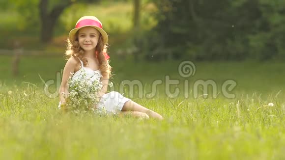 婴儿坐在一片草地上戴着一顶沙拉帆和帽子抱着野花和微笑慢动作视频的预览图