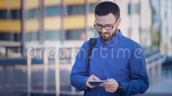 穿着蓝色衬衫和棕色裤子的长着大胡子肩上背着书包的英俊男子正在用他的手机打电话视频的预览图