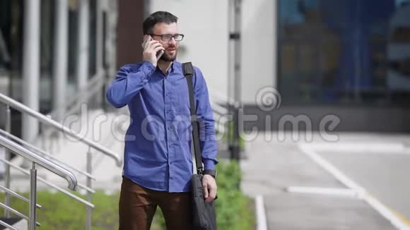 年轻英俊的胡须男穿着蓝色衬衫和棕色裤子站在户外打电话商人视频的预览图