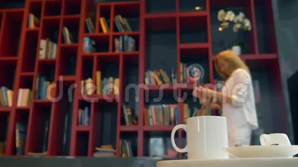 专注于一个杯子和一杯热饮料在图书馆的桌子上布鲁尔年轻的女人选择一本书阅读站在里面视频的预览图