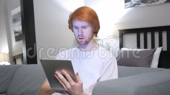 震惊好奇红发男子在卧室里用平板电脑视频的预览图