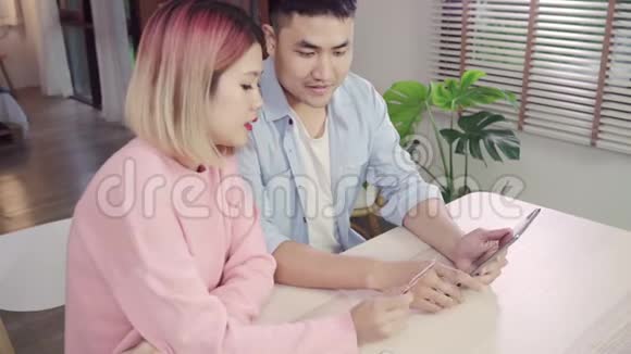 年轻的亚洲家庭夫妇使用平板电脑讨论新闻或做网上购物一起坐在办公桌上周末在家里视频的预览图