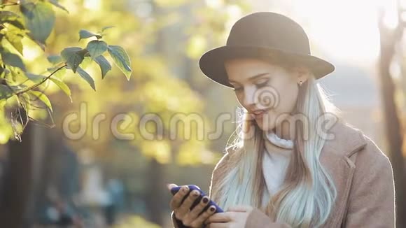 快乐的年轻女人穿着外套用智能手机站在秋天的公园里户外科技阳光视频的预览图