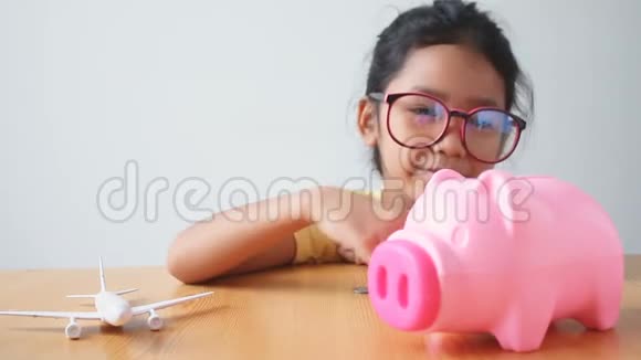 亚洲小女孩把硬币放进粉红色的储蓄罐里空中飞机比喻为旅行和运输概念省钱视频的预览图