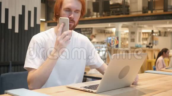 红头发胡子男在手机上说话坐在咖啡厅视频的预览图