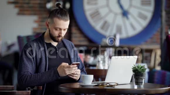 穿着黑色T恤和蓝色夹克的英俊留胡须的男人正坐在咖啡馆里用现代笔记本电脑和视频的预览图