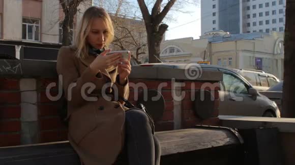 在市中心的夕阳下一个坐在长凳上的手机很时髦的女人视频的预览图