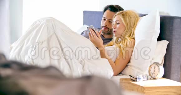 金发女人早上和男朋友在床上用智能手机恋爱中的情侣在床上醒来视频的预览图
