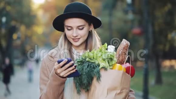 年轻漂亮的女人穿着时髦的外套站在秋天的公园里拿着一包产品用智能手机视频的预览图