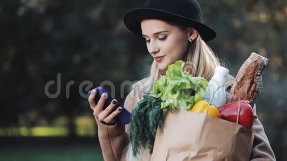 年轻漂亮的女人穿着时髦的外套站在秋天的公园里拿着一包产品用智能手机视频的预览图