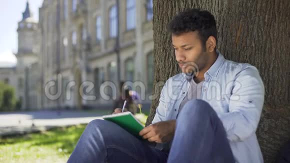 跨国公司的家伙坐在树下写着笔记本思考着作曲家视频的预览图