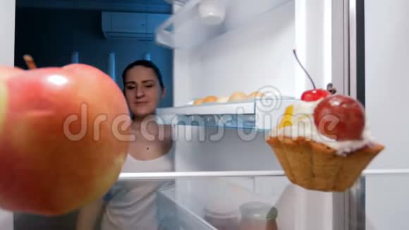 一个年轻微笑的女人晚上从冰箱里拿红苹果的镜头视频的预览图