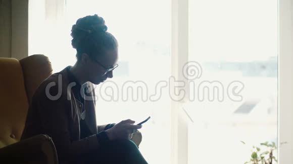 年轻漂亮的女人坐在靠近窗户的黄色椅子上用智能手机在网上聊天视频的预览图