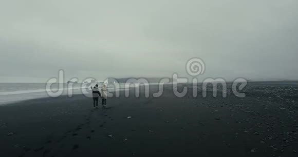 这对年轻夫妇穿着冰岛毛衣在冰岛黑色火山海滩的海边漫步视频的预览图