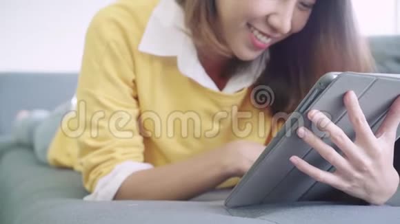 美丽的亚洲女性使用平板电脑观看视频而穿毛衣躺在沙发上的客厅在家里家庭生活方式妇女视频的预览图