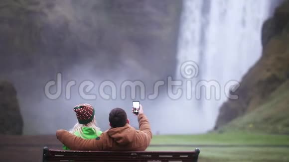 年轻夫妇坐在长凳上在智能手机上自拍照片的背面冰岛的斯科加沃斯瀑布视频的预览图