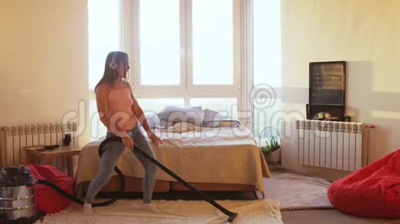 美丽的女人正在家里用现代吸尘器打扫地板用耳机听音乐视频的预览图