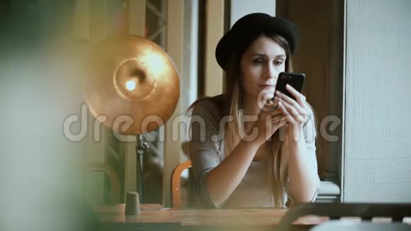 戴着智能手机和在咖啡馆使用无线技术的年轻美女帽子的肖像视频的预览图