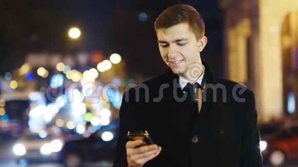 一个年轻人看着手机屏幕晚上站在城市的背景上视频的预览图