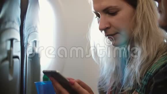 年轻漂亮的金发女人坐在飞机的窗户附近用智能手机两个人一起旅行视频的预览图