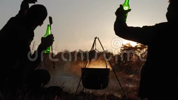 快乐的两个旅游朋友在日落时坐在森林的篝火旁取暖团体旅行者野餐视频的预览图